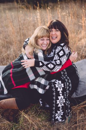 Two friends share joyful moment wrapped in patterned blankets in autumn field.