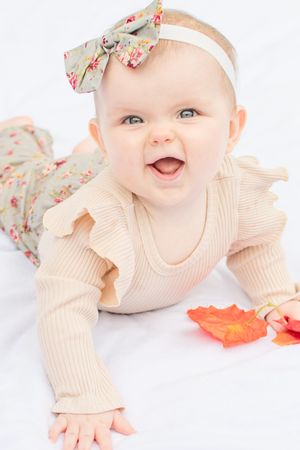 A baby in a cream ribbed outfit and floral bow headband lies on their tummy reaching for autumn leaves.