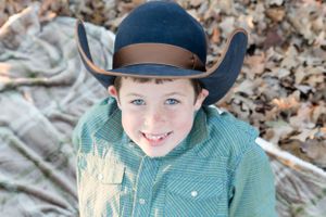Young person wearing navy cowboy hat smiling outdoors among fallen leaves.