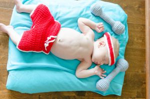 Baby wearing red and gray crochet superhero outfit lying on turquoise blanket.
