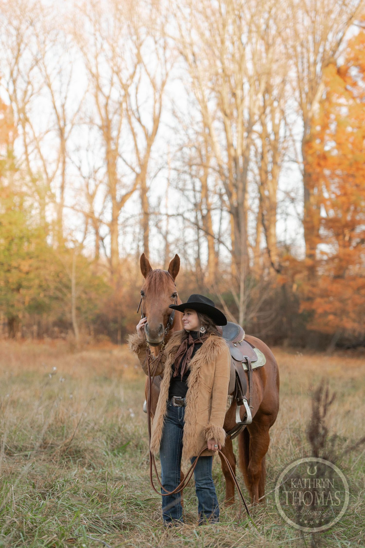 A chestnut horse with saddle stands beside a figure in western wear against autumn trees.
