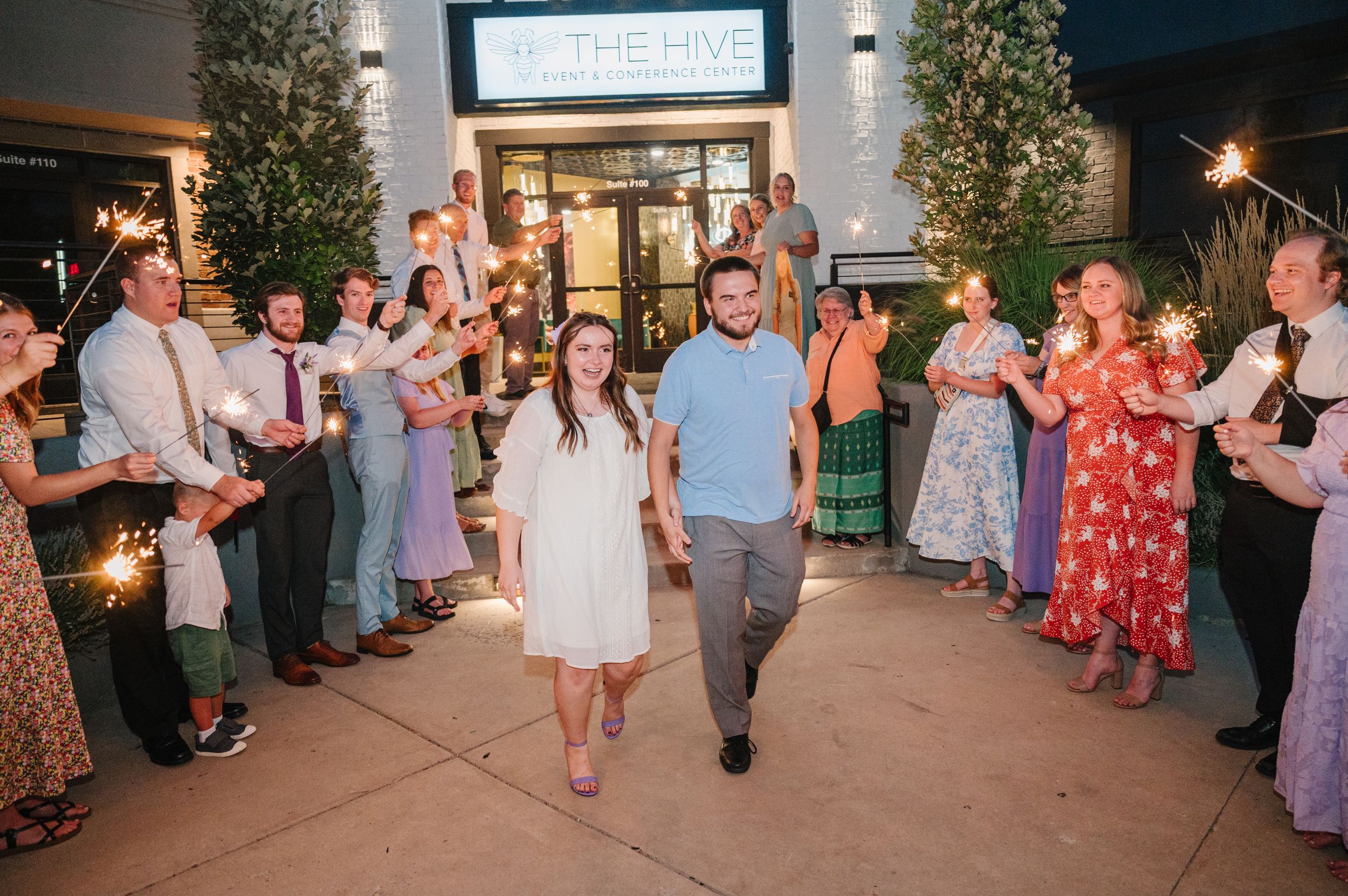 A couple walks through sparkler exit at nighttime wedding celebration as guests line pathway outside venue entrance.