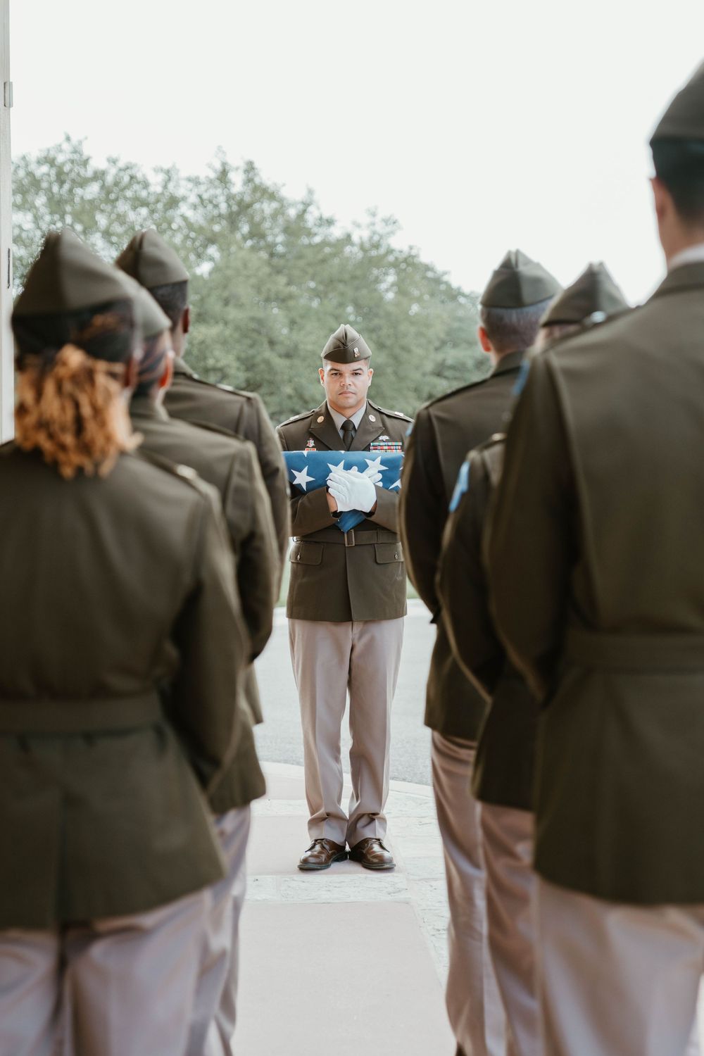Military ceremony with soldier holding folded American flag, flanked by uniformed cadets standing at attention outdoors.