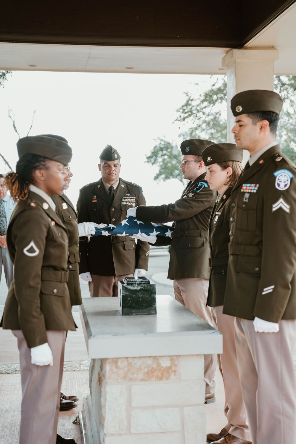 Military honor guard soldiers in olive uniforms performing a flag folding ceremony at a memorial service.