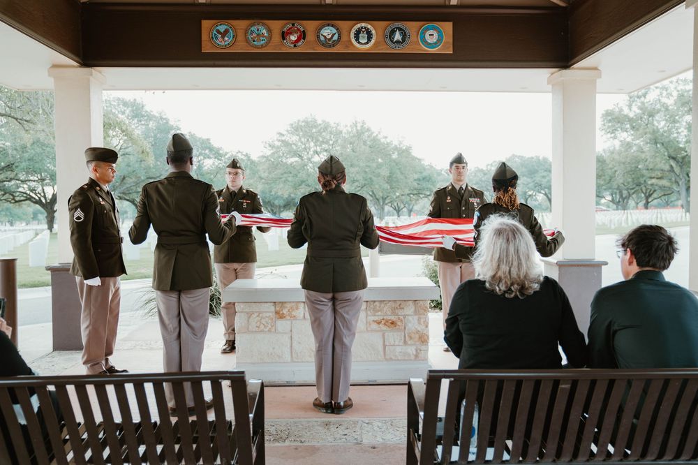 Military honor guard performing a flag folding ceremony at a memorial pavilion with seated attendees.