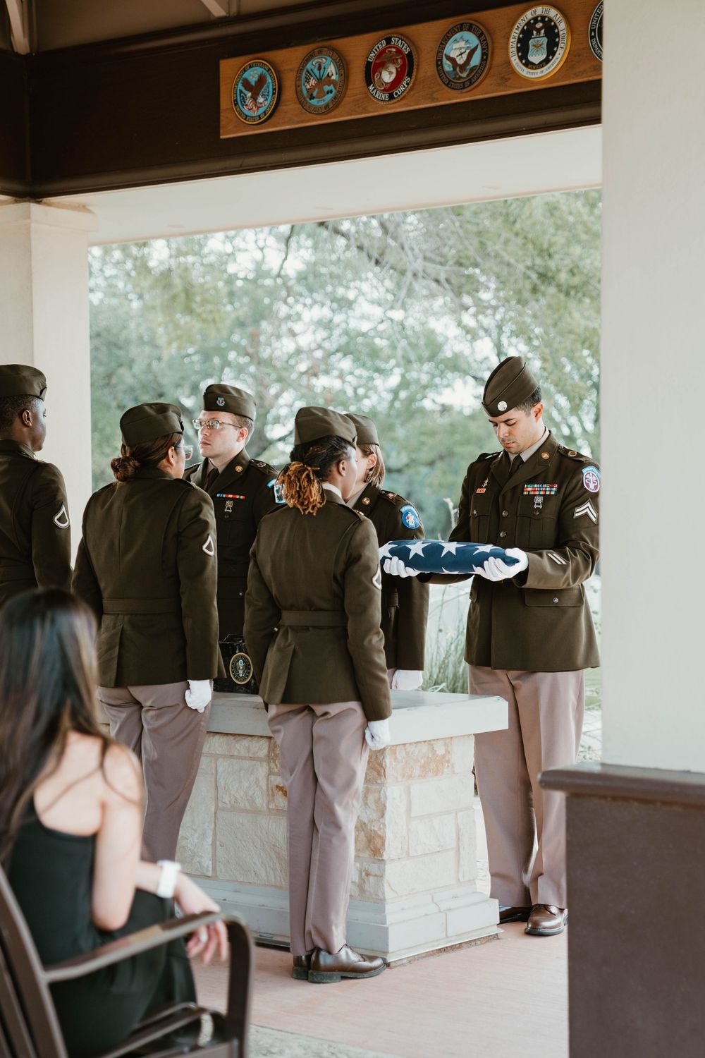 Military honor guard folding flag at a funeral ceremony, soldiers in dress uniforms at an outdoor memorial pavilion.