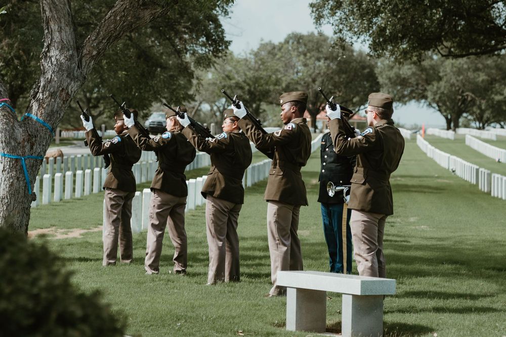 Military honor guard firing rifles in salute at a cemetery burial ceremony among green trees and white headstones.