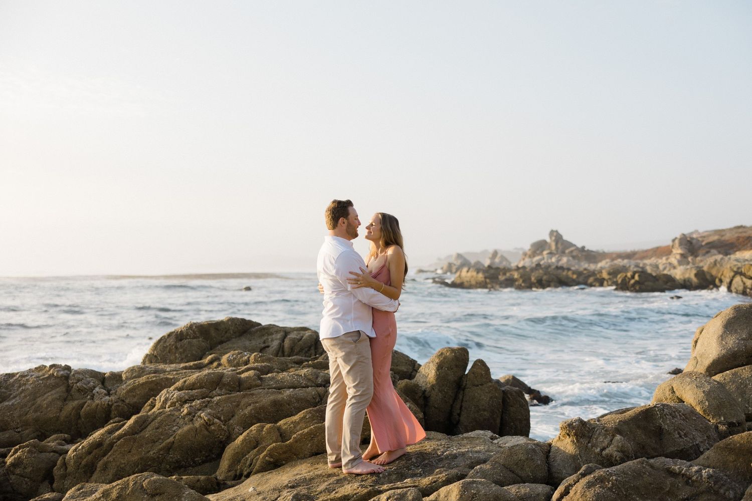 A couple embraces on rocky coastal cliffs at sunset while waves crash against the California shoreline.