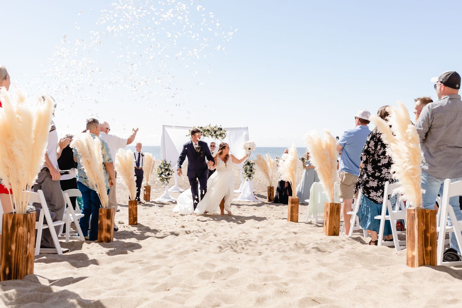 Joyful beach wedding ceremony with pampass grass decorations and guests seated in white chairs along sandy aisle.