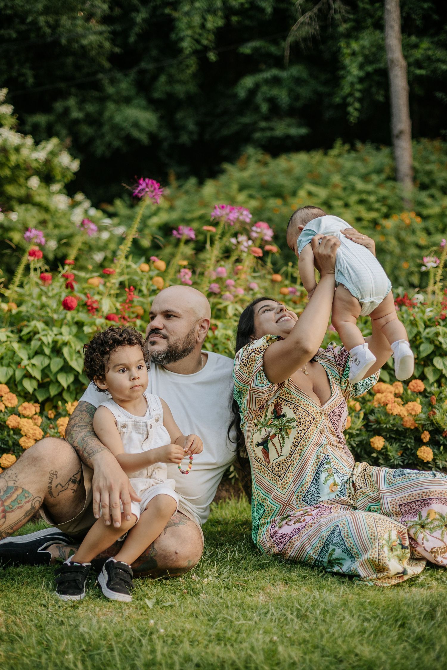 family of 4, sitting in the grass full of flowers on the background, the older one looking away and mom raising up her baby