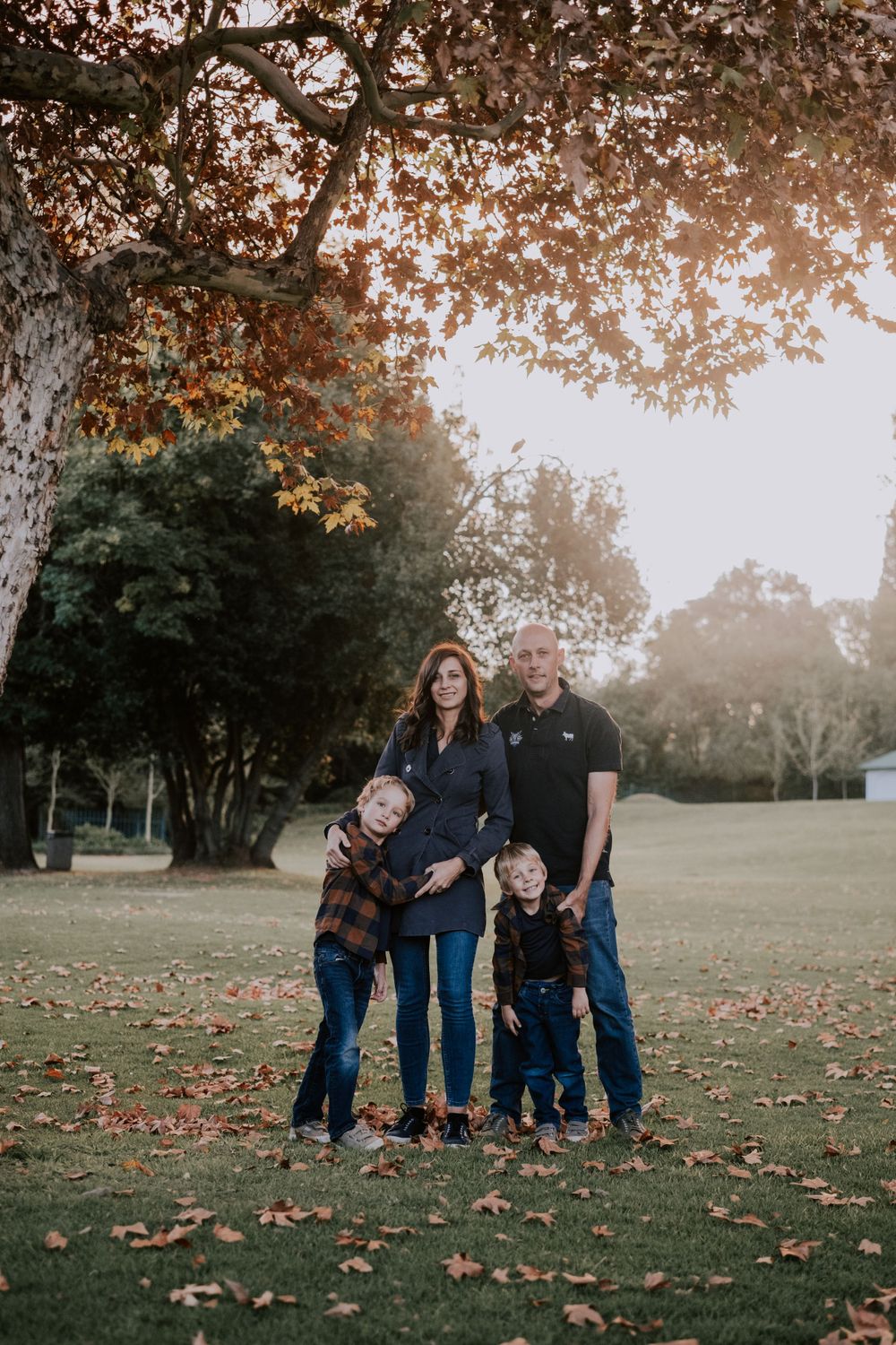 A family stands together under a large tree with golden autumn leaves in a park setting at sunset.