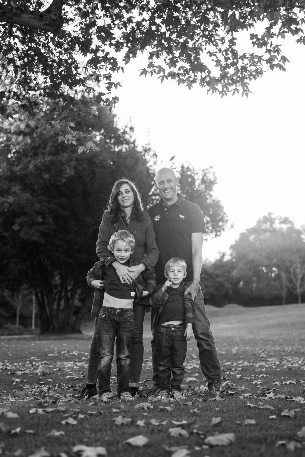 A family of four poses together on fallen autumn leaves in a park setting, captured in black and white.