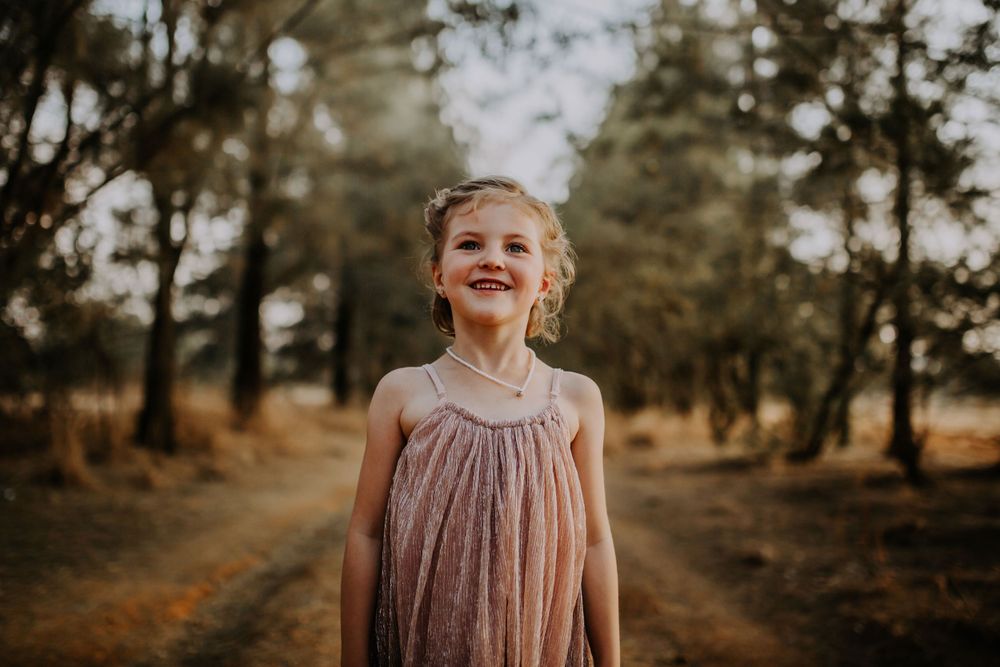Young child in dusty pink dress smiles brightly on forest path at golden hour.