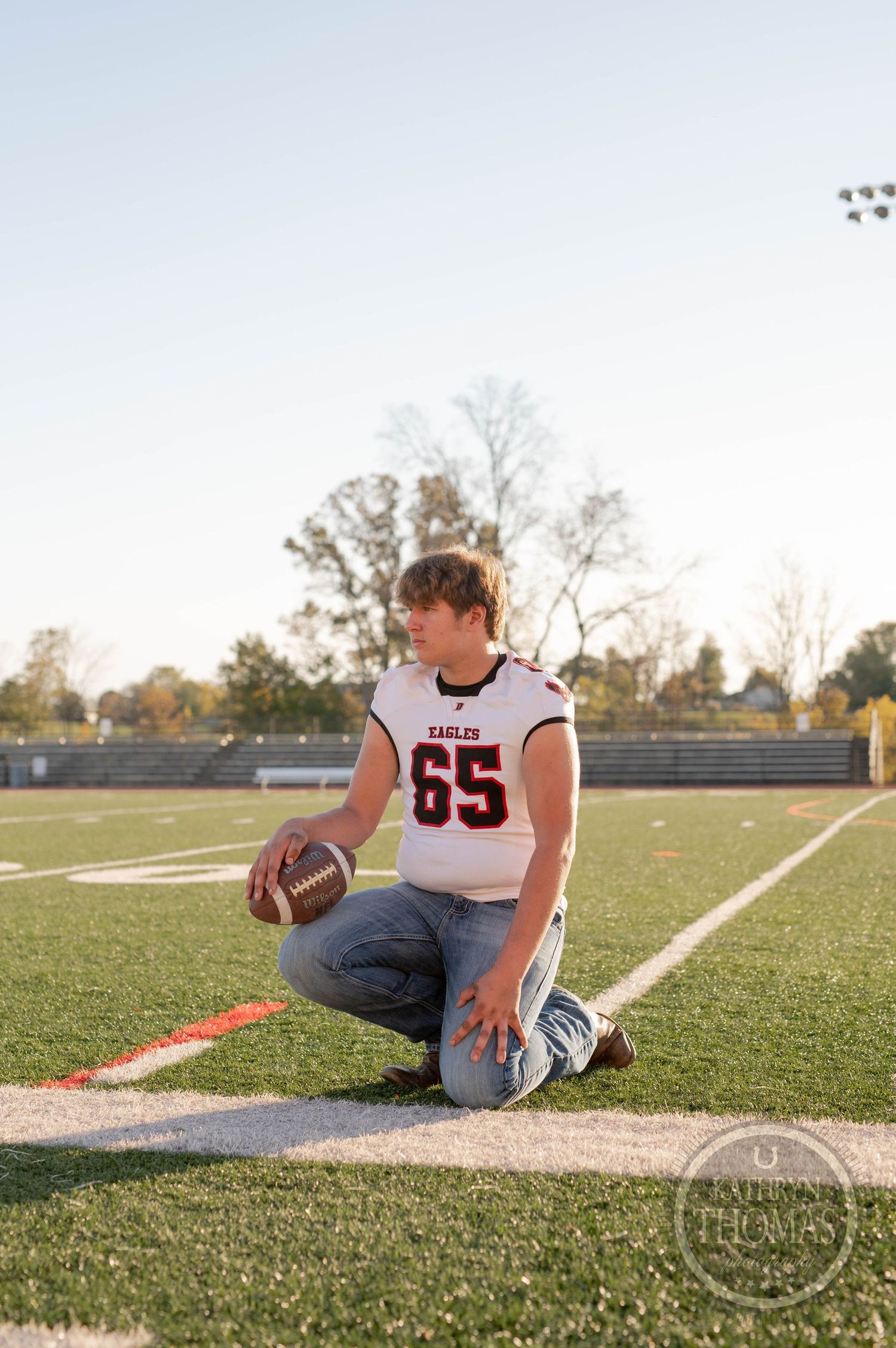 Athlete in white uniform kneels with football on field sideline at dusk.
