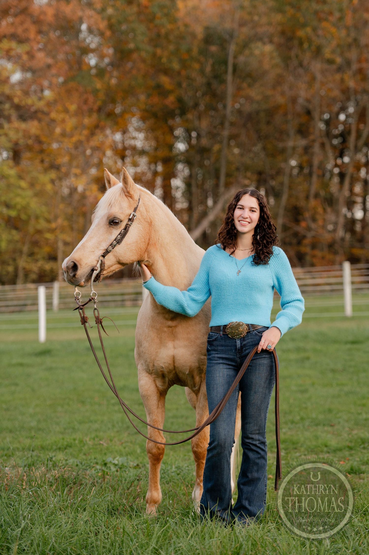 A cream-colored horse stands calmly in a fenced pasture surrounded by fall trees.