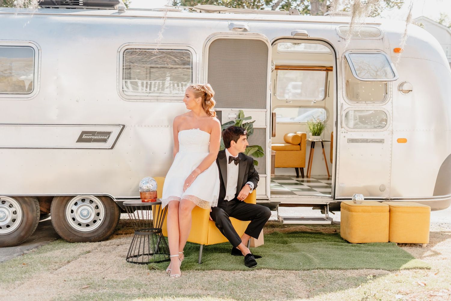 A couple in formal attire poses outside a vintage Airstream trailer with yellow suitcases on a sunny day.