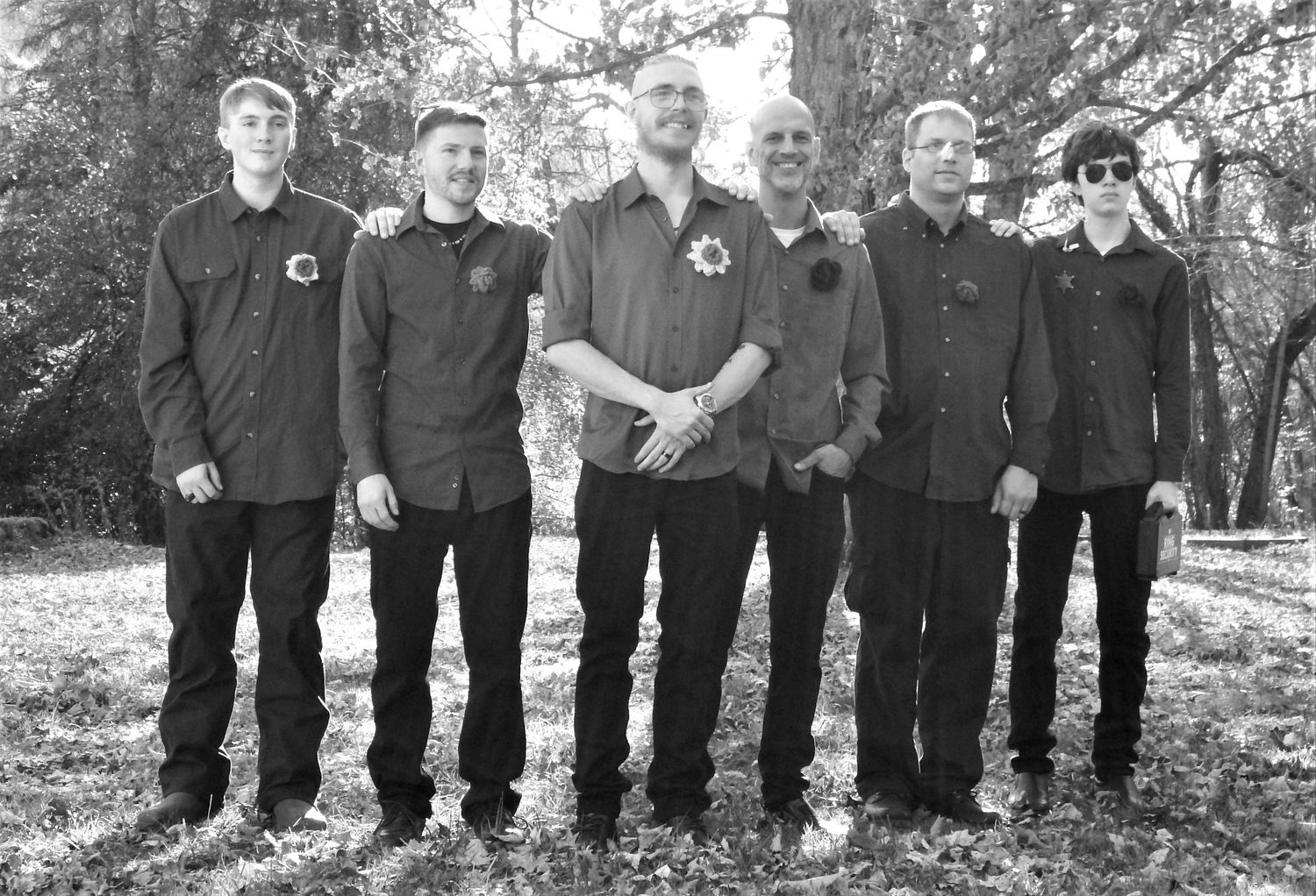 A group stands in a line wearing matching dark outfits in a wooded setting.