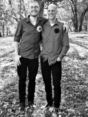Two people in dark clothing stand together on fallen leaves in a forest.