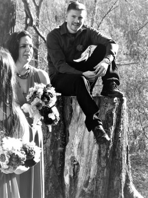 Black and white wedding photo of a couple and their dog posing playfully near a tree in a forest setting.
