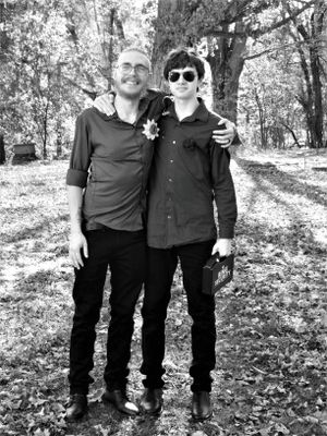 Group portrait of wedding attendees in coordinated black outfits standing in a wooded outdoor setting.