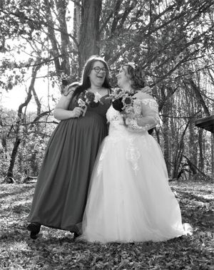 Two people in formal attire pose together in a wooded outdoor setting, one in dark dress and one in white gown.