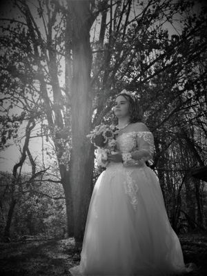 An ethereal black and white portrait captures someone in a flowing white gown among sunlit trees.