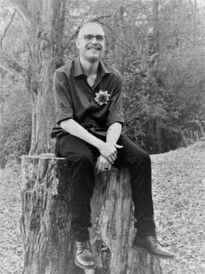 A relaxed outdoor portrait shows someone seated on a fallen tree trunk in casual dark attire.