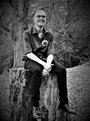 Black and white photo of someone sitting on a tree stump in a natural woodland setting.