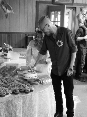 A couple examines their wedding cake and cupcakes displayed on a white tablecloth at their reception.