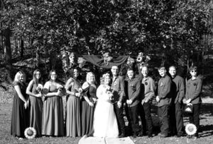 A wedding party poses outdoors with bridesmaids in dark dresses and groomsmen in casual attire.