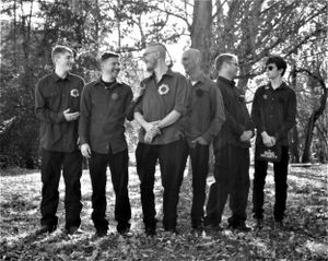 A line of groomsmen in black attire pose together outdoors among trees.