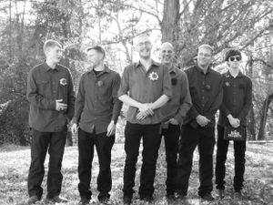 Wedding party members in dark attire stand in a line against a natural outdoor backdrop.