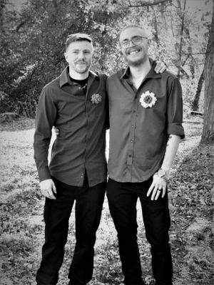 Groomsmen in black shirts pose together for formal photos in a wooded setting.