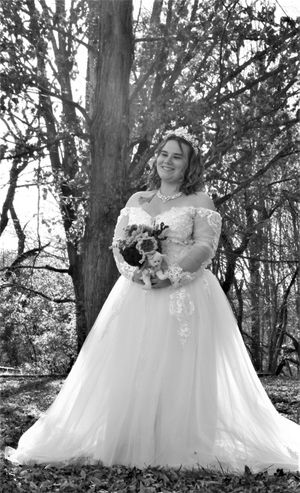 A bride in an ornate white dress holds a bouquet while posing among trees.