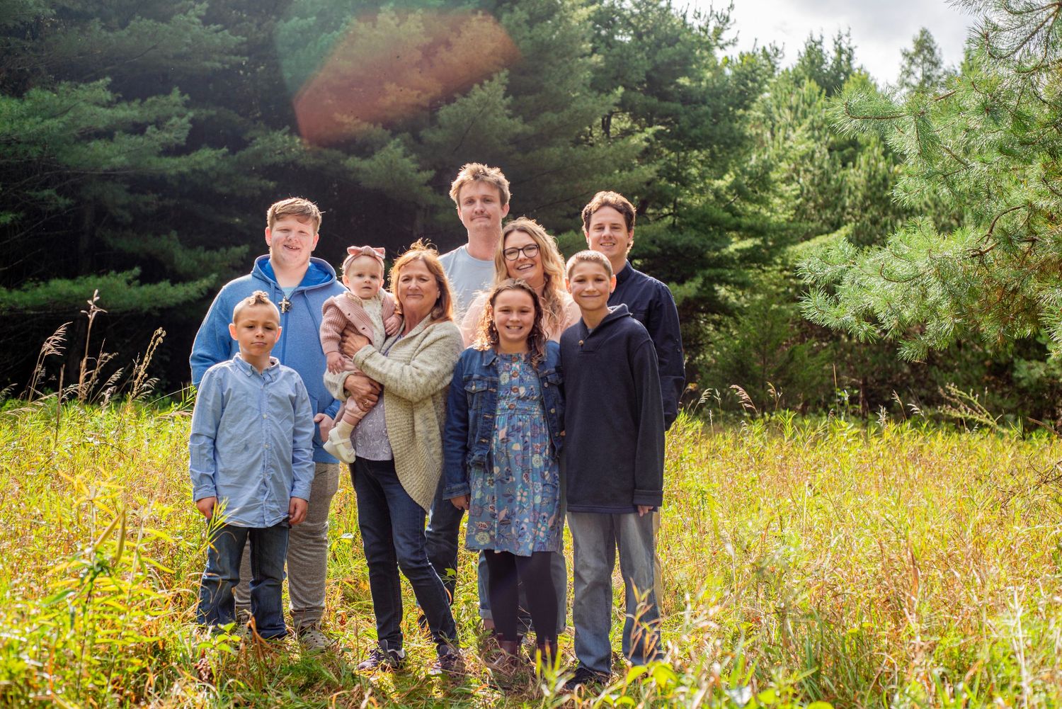 A group stands together smiling in a sunny meadow with evergreen trees in the background on a bright summer day.