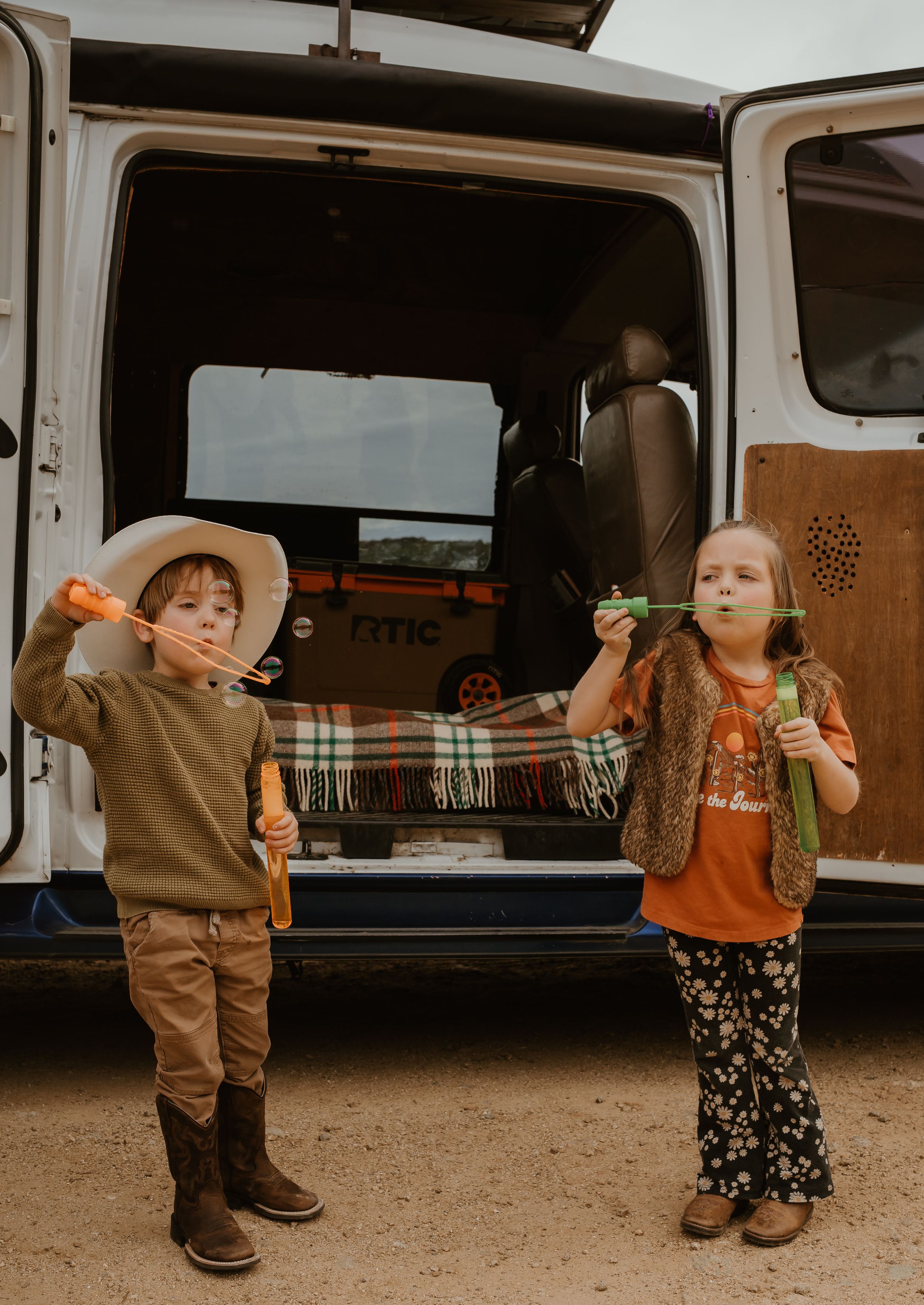 Two children stand beside a vintage van blowing bubbles while wearing casual clothes and western-inspired outfits.