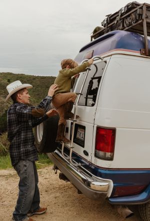 Two people work together to lift a large brown dog into the back of a white van on a dirt road in a rural area.