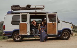 Camper van with open sliding door parked in nature with travelers sharing a casual moment inside.