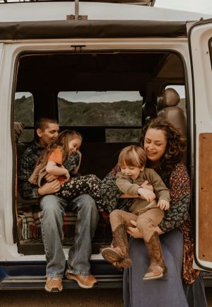 A family sits together in the open trunk of a van with a scenic mountain view in the background.