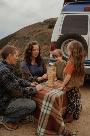 A family plays Jenga together on a plaid blanket while camping next to their SUV in the mountains.