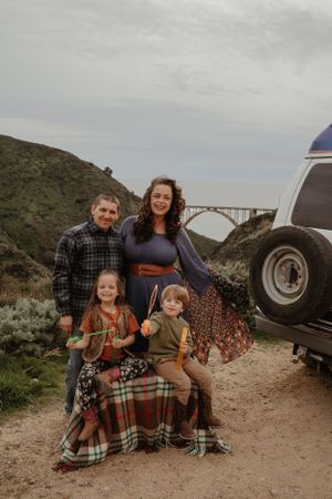 Family in plaid sits against classic RV on a mountain trail with scenic bridge view in the background.