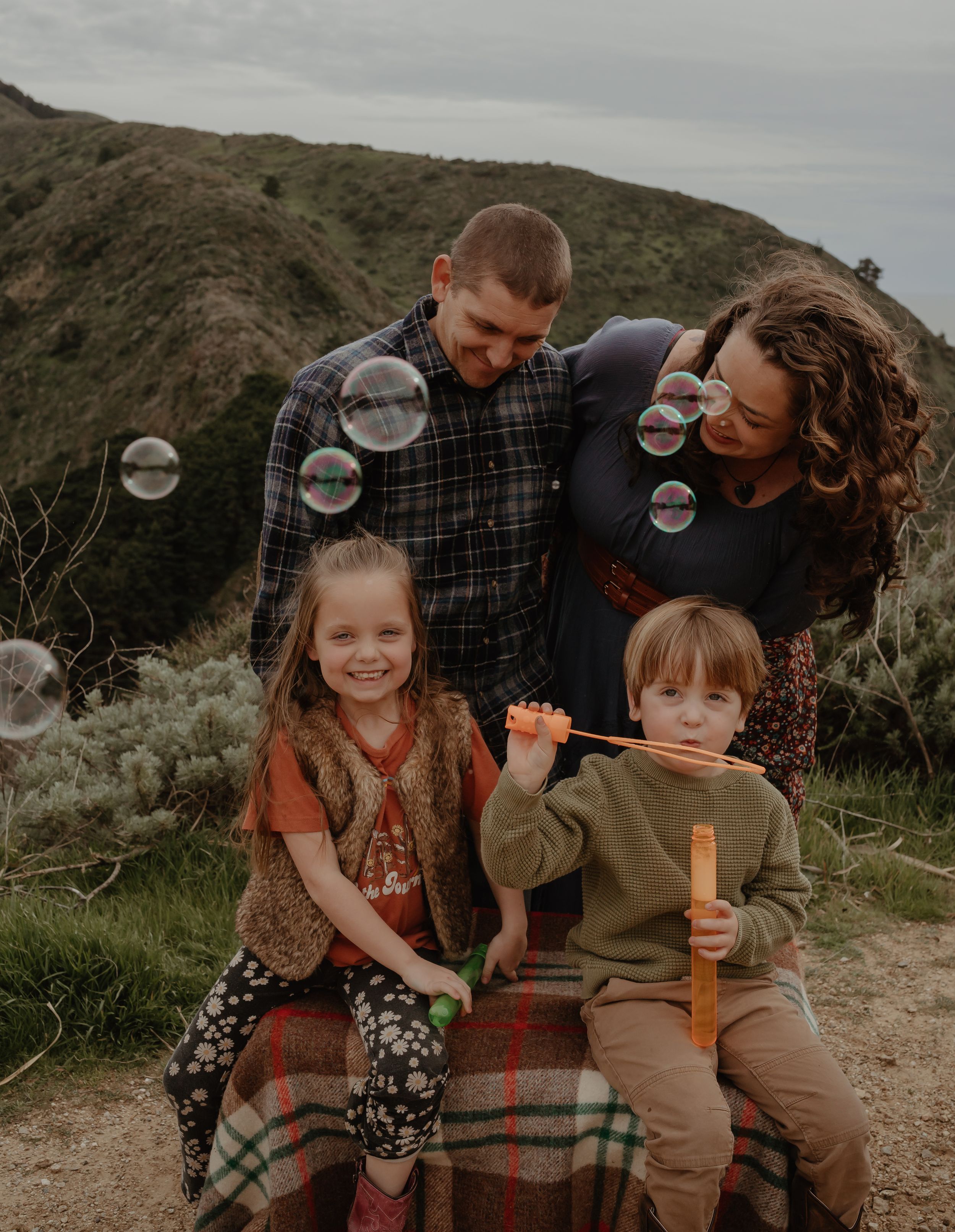 A family blowing bubbles together while sitting on a blanket on a hillside path during an overcast day.