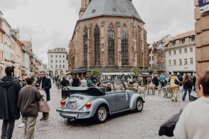 Hellblauer Oldtimer, der über den Markplatz in Heidelberg fährt. Im Hintergrund sieht man eine Kirche, Leute laufen über den Platz.