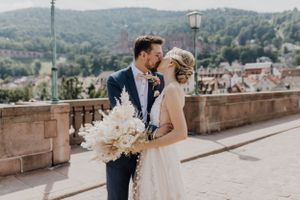 Küssendes Brautpaar in Heidelberg auf der alten Brücke. Im Hintergrund erahnt man das Schloss, die Braut trägt einen hellen Brautstrauß.