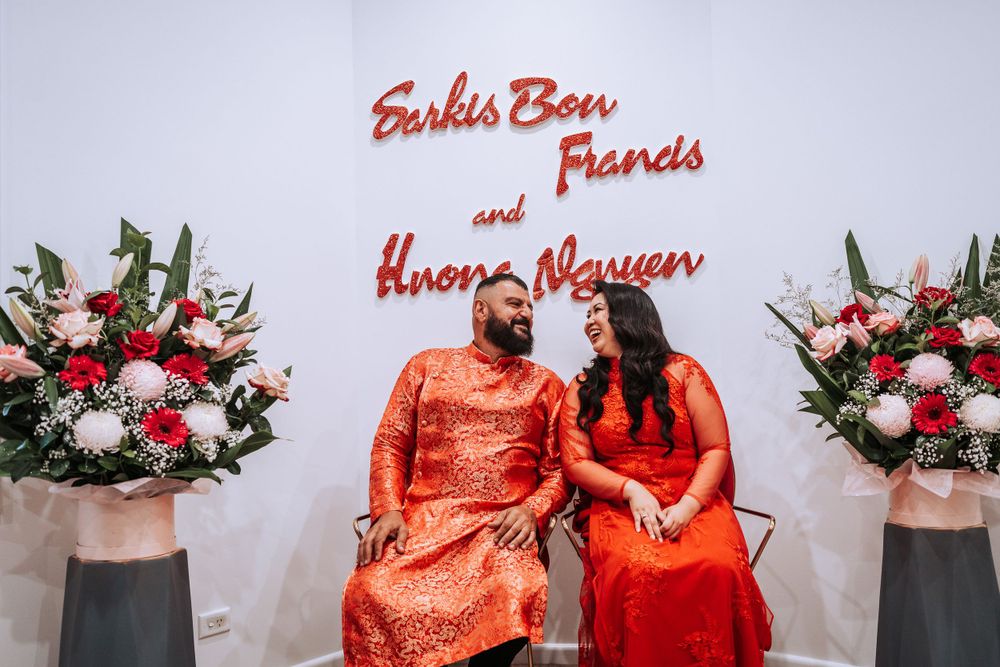 Two people in matching coral outfits sit together against a wall with red text and floral arrangements.