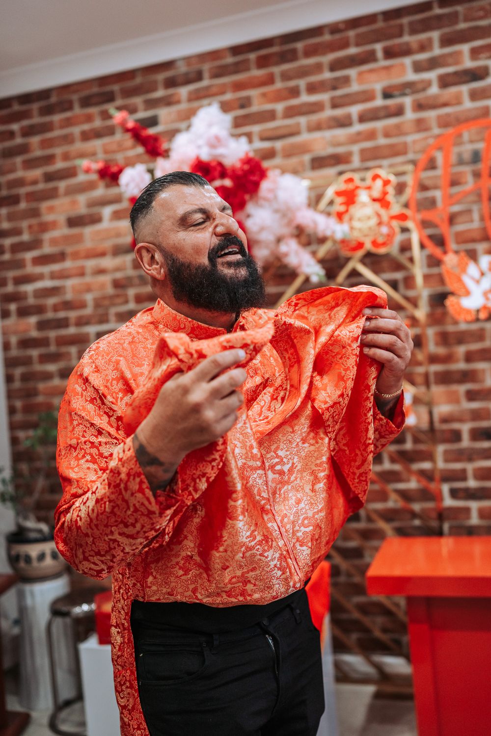 Man wearing orange traditional patterned shirt laughs joyfully while holding fabric against brick wall background.