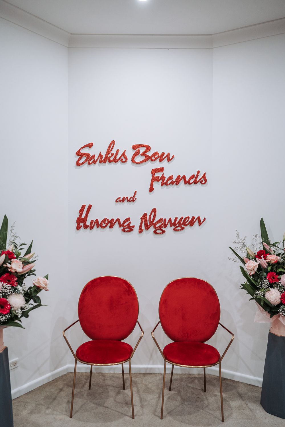 Modern event space setup with two red velvet chairs and floral arrangements beneath red lettered wall signage.
