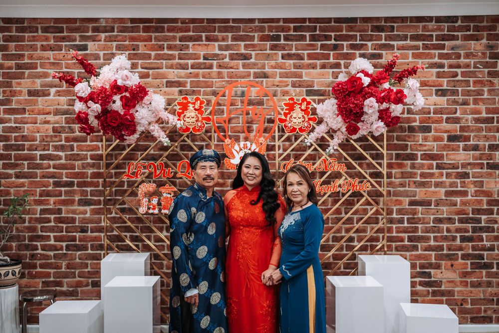Three people pose in front of a brick wall decorated with red and pink balloon arrangements for Lunar New Year.