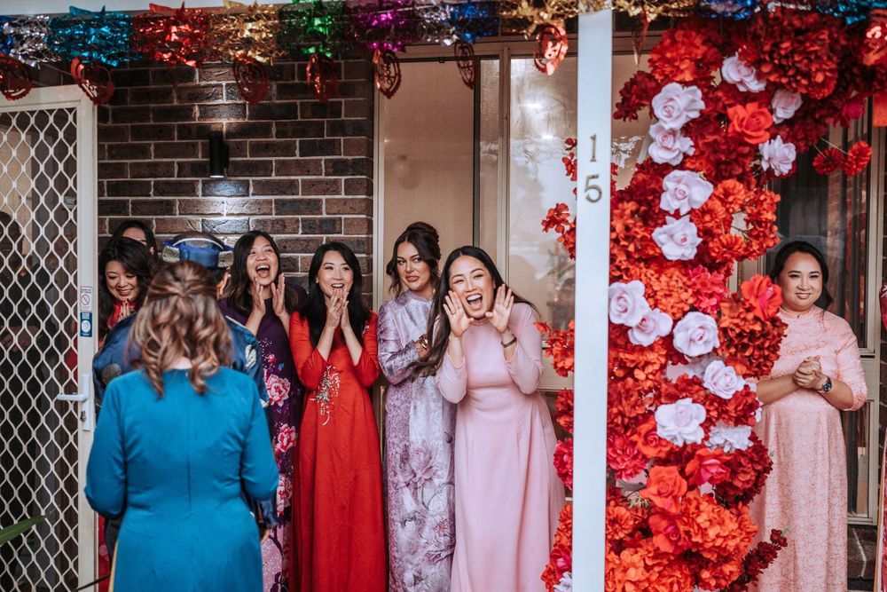 Wedding party gathered under colorful floral decorations and string lights against a brick wall background.