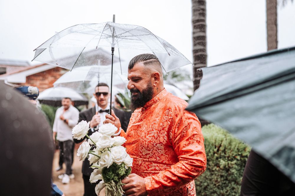 Someone in a bright coral outfit holds a white floral bouquet while standing under a clear umbrella in the rain.